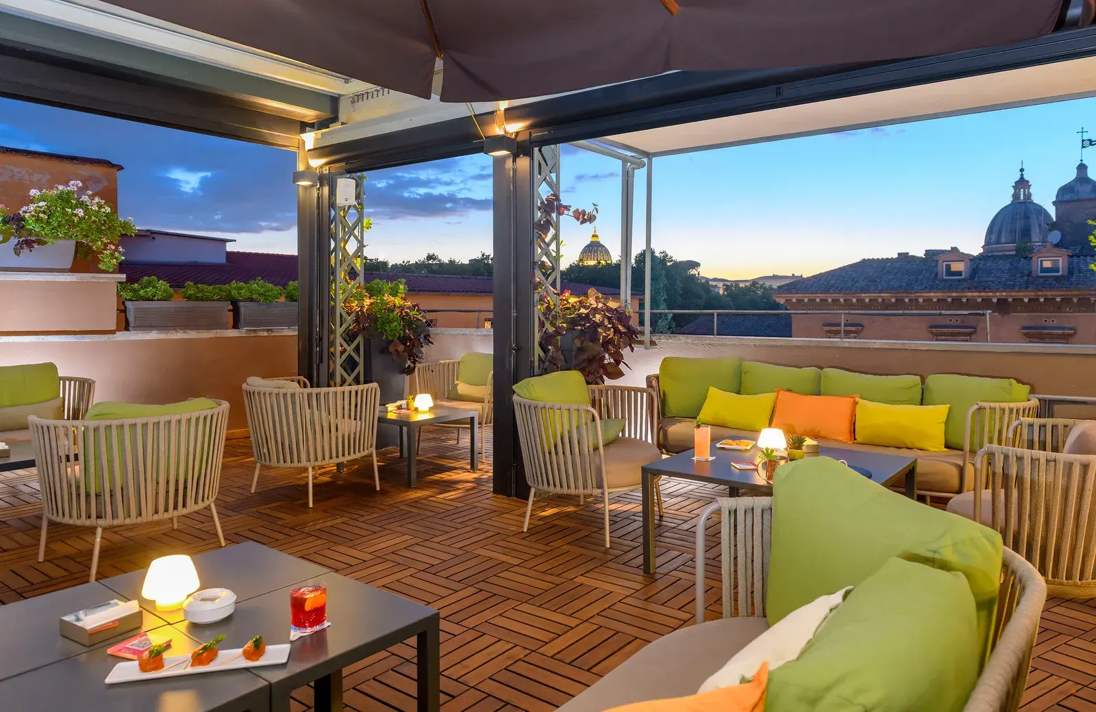 Rooftop terrace with lounge seating and soft lighting at dusk, with Roman domes in the distance